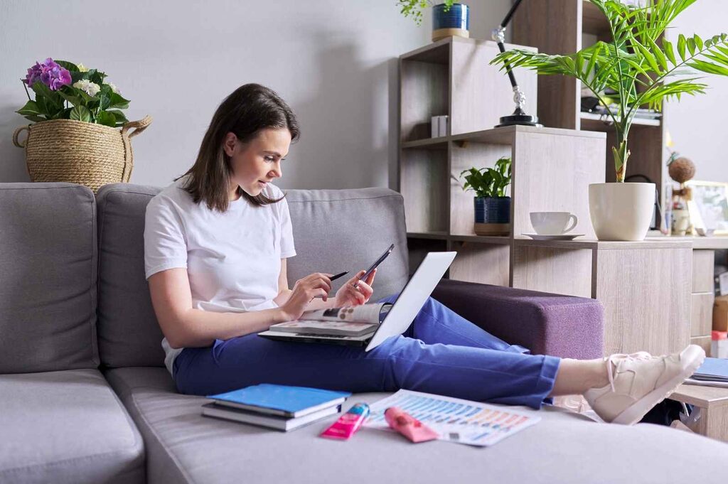 Woman is sitting on her living room couch with a laptop resting on her tights and holding a mobile phone, researching how to pick a blog niche to start an online business.