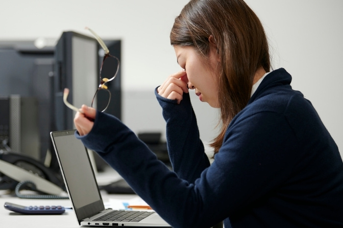 An Asian woman rubs her tired eyes with one hand and holds her glasses with the other. She sits in front of a laptop, feeling lazy and lacking the motivation to return to work.