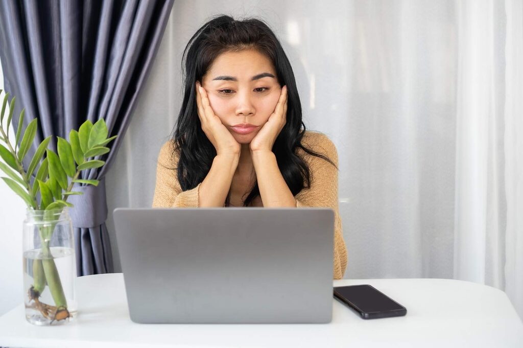 A woman, her chin resting on her palms, looks wearily at a laptop. The image illustrates the importance of training your brain to stop being lazy, rewarding yourself for being productive.