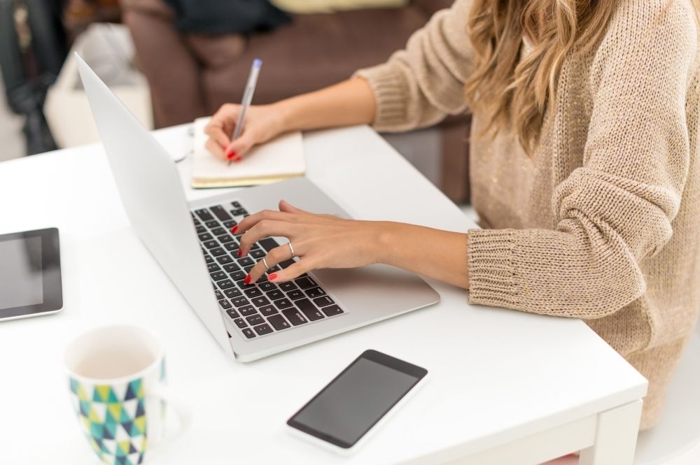 Woman typing on a laptop with one hand and writing on a notebook with the other.
