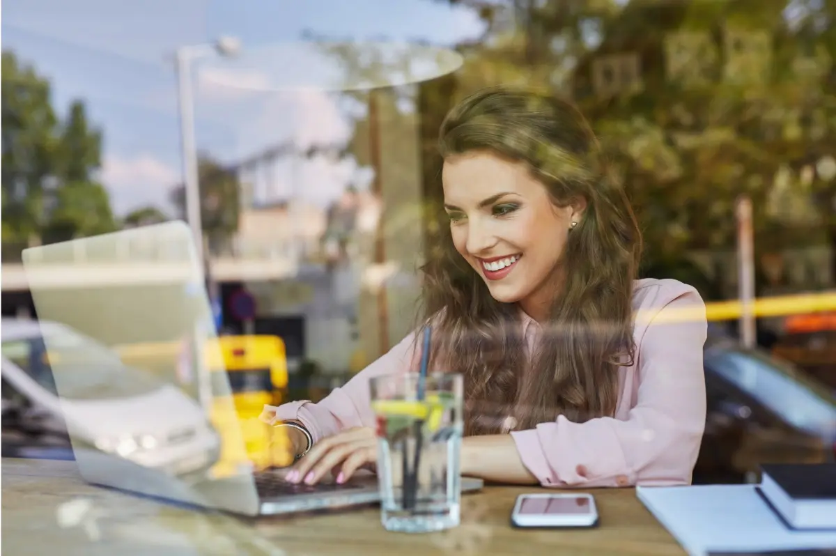 Photo of a woman, seen through the glass wall of a coffee shop, smiling confidently and typing on a notebook.