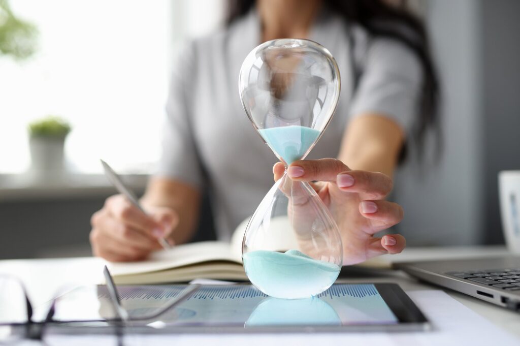 Close-up of a woman's hand holding an hourglass. In the blurred background, the woman is writing something on a notebook in front of a laptop.