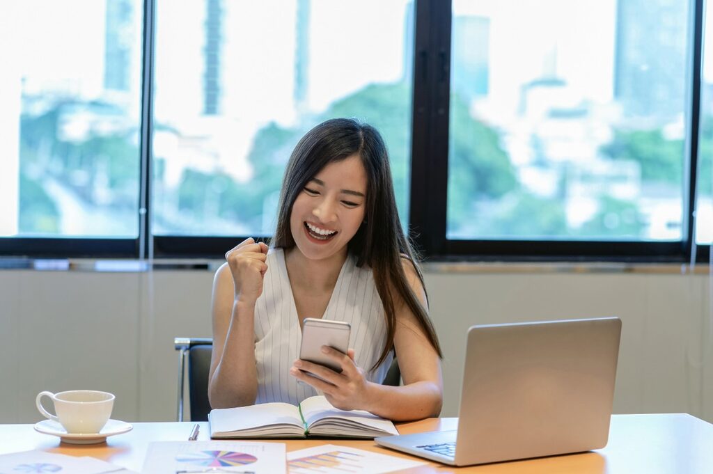 Young Asian female entrepreneur commemorating something she's checking on her phone in front of an office desk with a laptop.