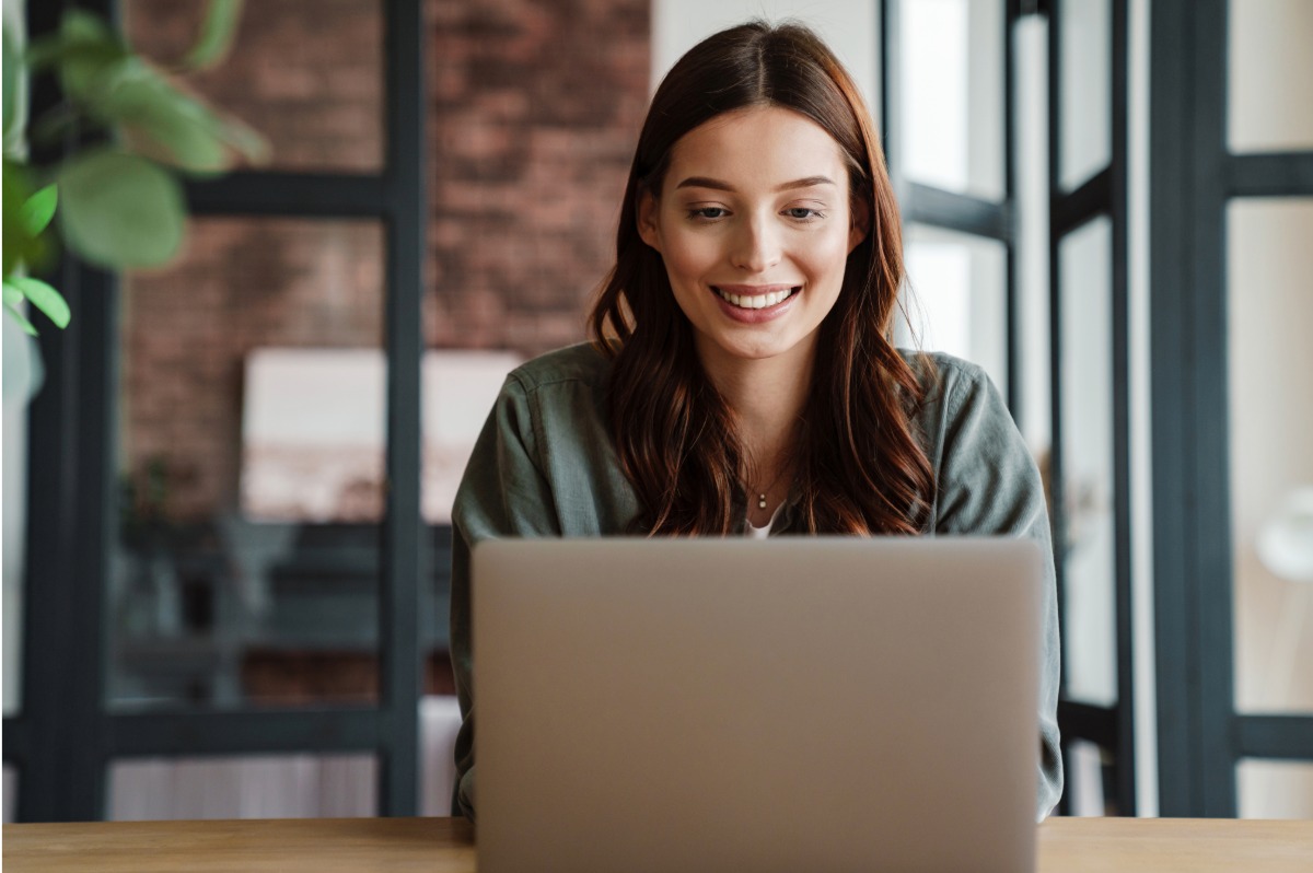 Young woman with chestnut hair color, wearing green and smiling at a laptop.