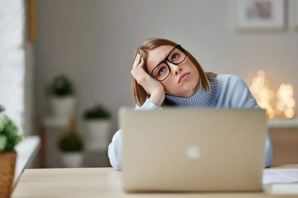 A woman with short straight hair, wearing a light blue sweater and glasses. She has her face resting on one hand and her elbow on the table. She appears bored and grumpy in front of a laptop.