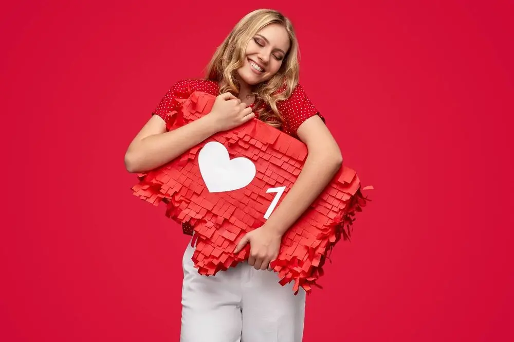 A blonde woman hugging a red sign that simulates the Instagram heart-shaped like bar, wearing a red t-shirt in front of a red background.