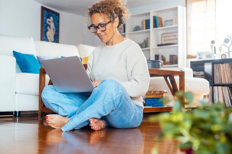 Woman sitting on the floor of a living room, wearing glasses and confidently typing on a grey laptop.