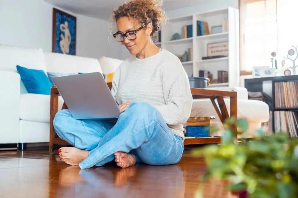 Woman sitting on the floor of a living room, wearing glasses and confidently typing on a grey laptop.