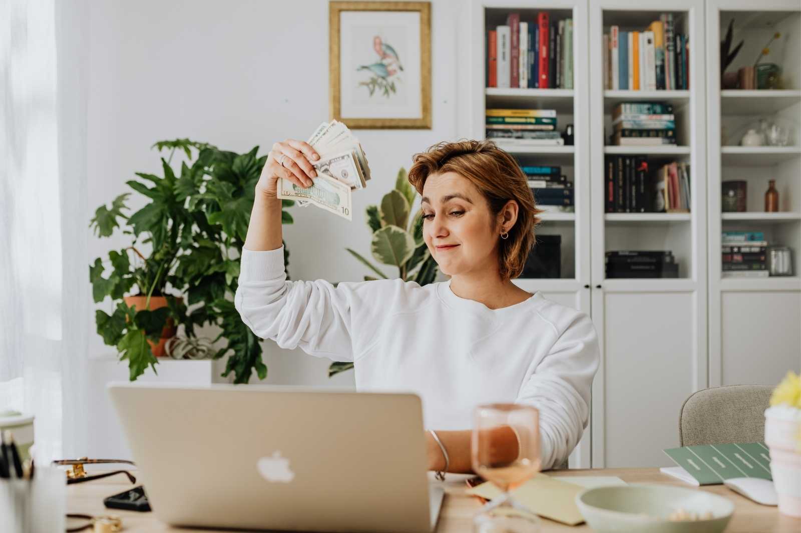 An adult woman, at her home office, holds a bunch of dollars in front of her laptop camera. The image illustrates profitable small business ideas from home with low investment.