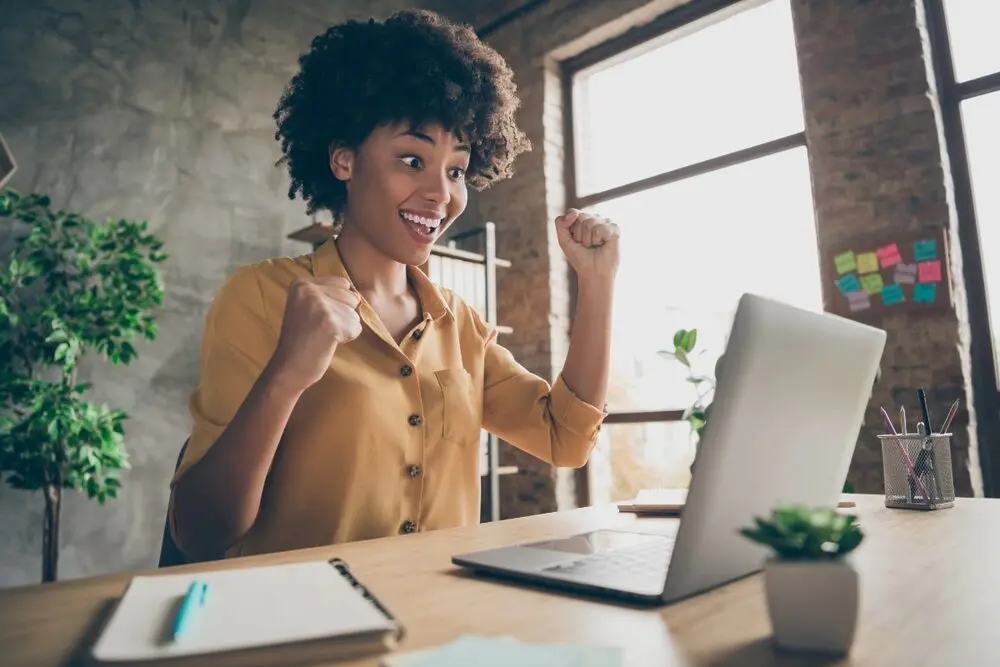 A black woman wearing a yellow cardigan is celebrating while reading something on her laptop screen.