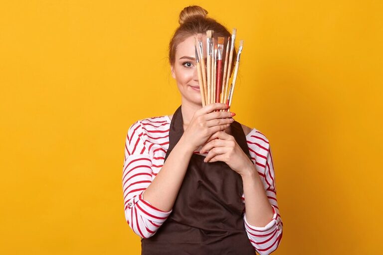 A red-haired woman with her hair tied in a bun, wearing an art apron and holding paintbrushes in front of her face. She is in front of a yellow background.