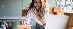 Young Asian entrepreneur owner talking on phone at workplace at home. The image illustrates the subject: "How Entrepreneurs Can Use Adobe Express to Build Smarter Marketing."