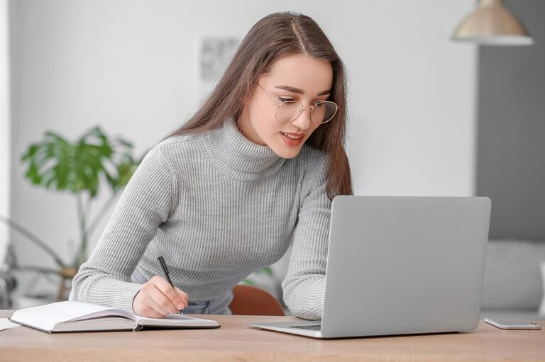 Woman wearing glasses and confidently looking at a laptop screen while taking handwritten notes on a notebook.