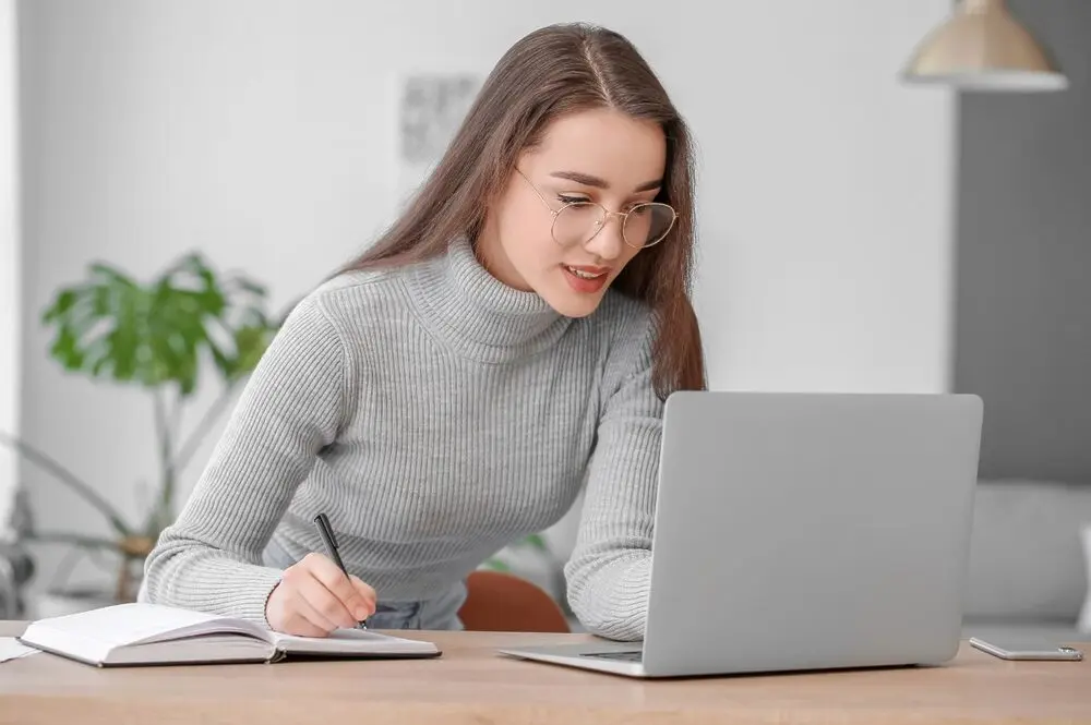 Woman wearing glasses and confidently looking at a laptop screen while taking handwritten notes on a notebook.