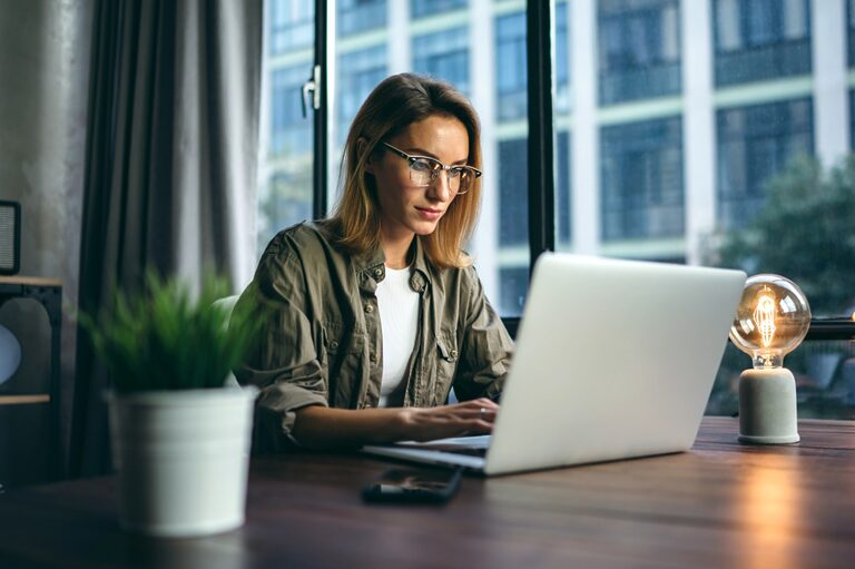 Blonde woman working on a laptop in front of a big office window.