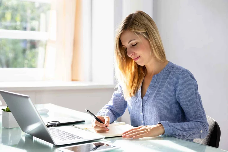 A blonde woman writing on a paper, in front of her laptop, and working from home.