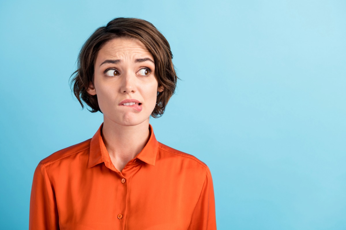 A woman with short hair and an orange blouse, biting her lower lip in apprehension.