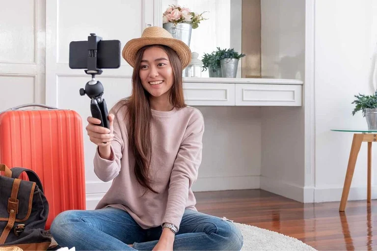 An Asian woman wearing a hat is sitting on the ground, holding a stand with a cell phone that she is using to film herself.
