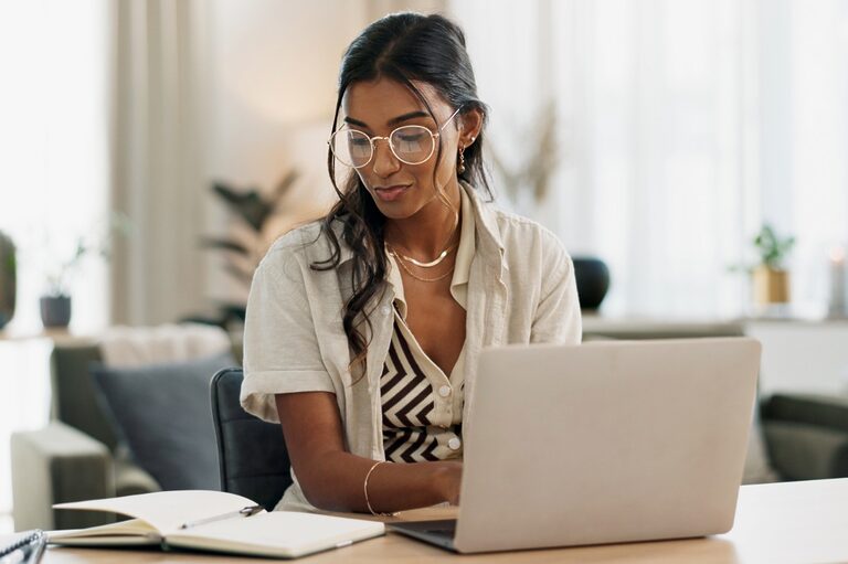Woman of color working with her laptop in her living room, checking notes on a notebook.
