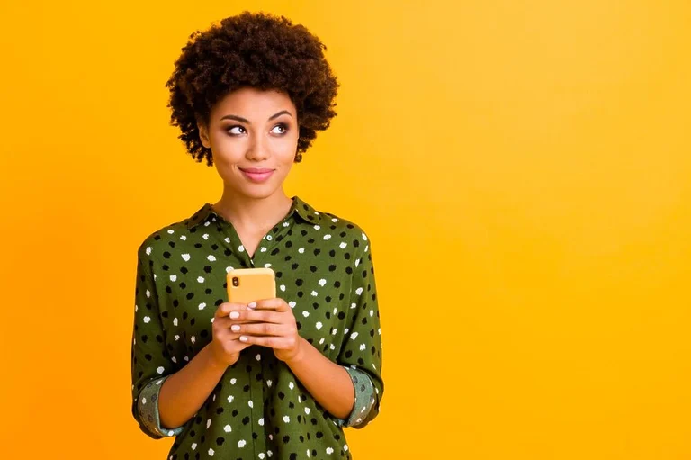 Woman wearing a militar green shirt, holding a yellow smartphone in front of a yellow wall.