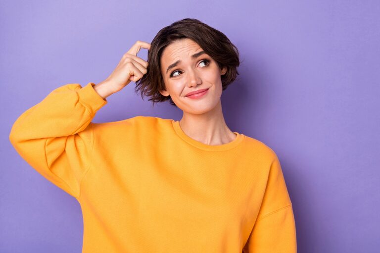 A woman scratches her head, expressing doubt. She is wearing a yellow blouse against a lilac background.