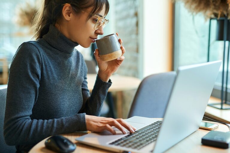 Woman wearing glasses and a sweater, drinking from a mug she's holding the left hand and typing on a laptop with a right hand.