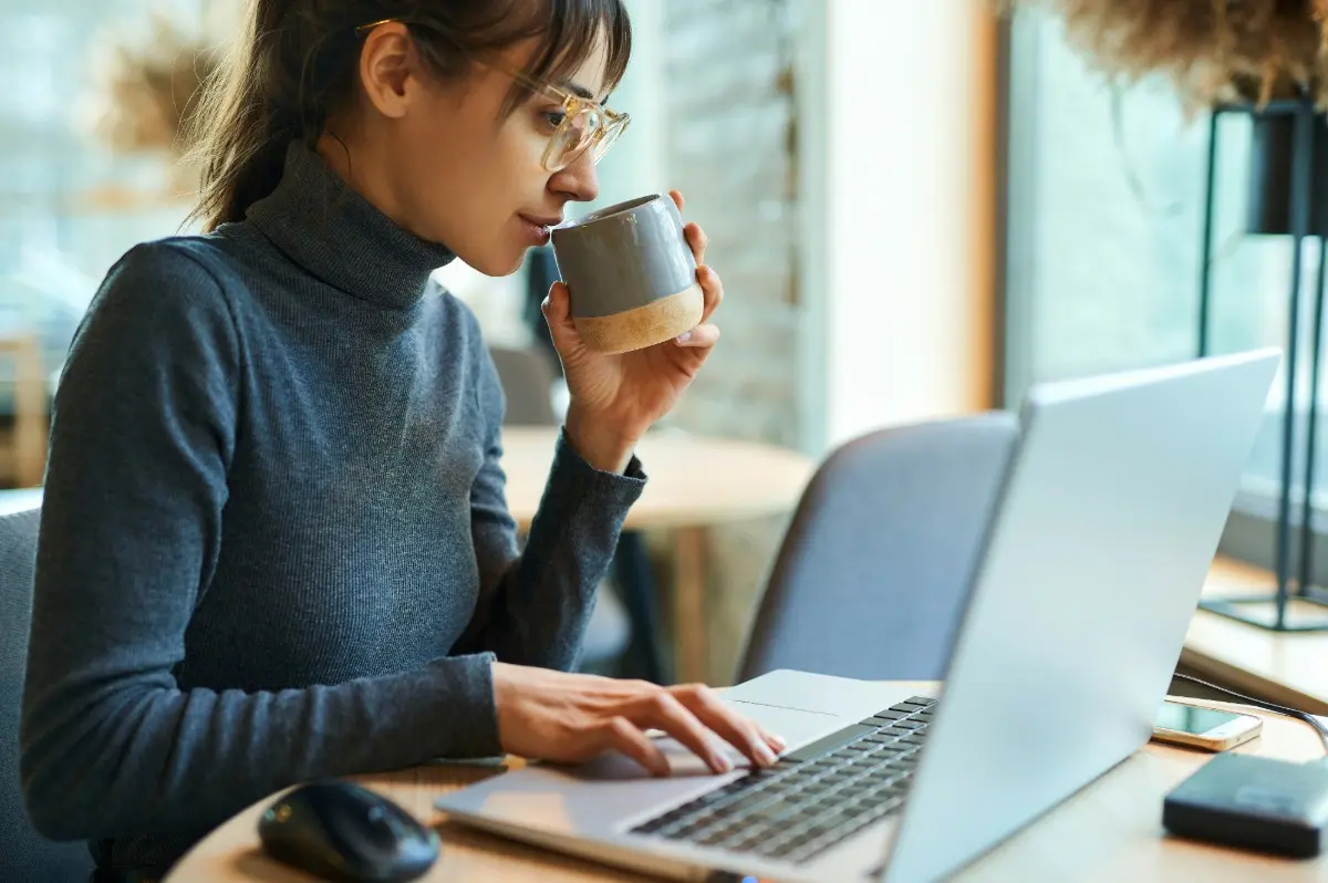 Woman wearing glasses and a sweater, drinking from a mug she's holding the left hand and typing on a laptop with a right hand.