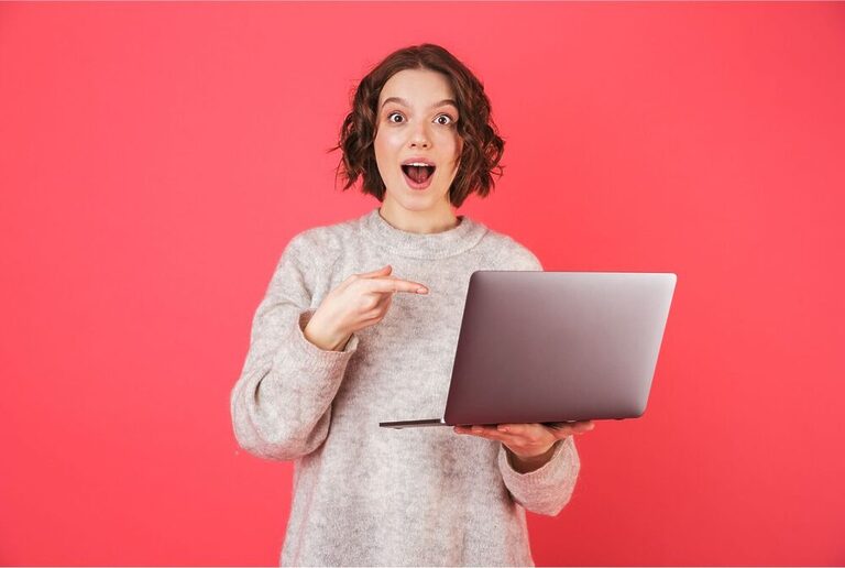 Short-haired woman smiling assertively while holding a laptop with one hand and pointing to it with the other one. She wears a grey sweater and has a blank reddish background behind her.