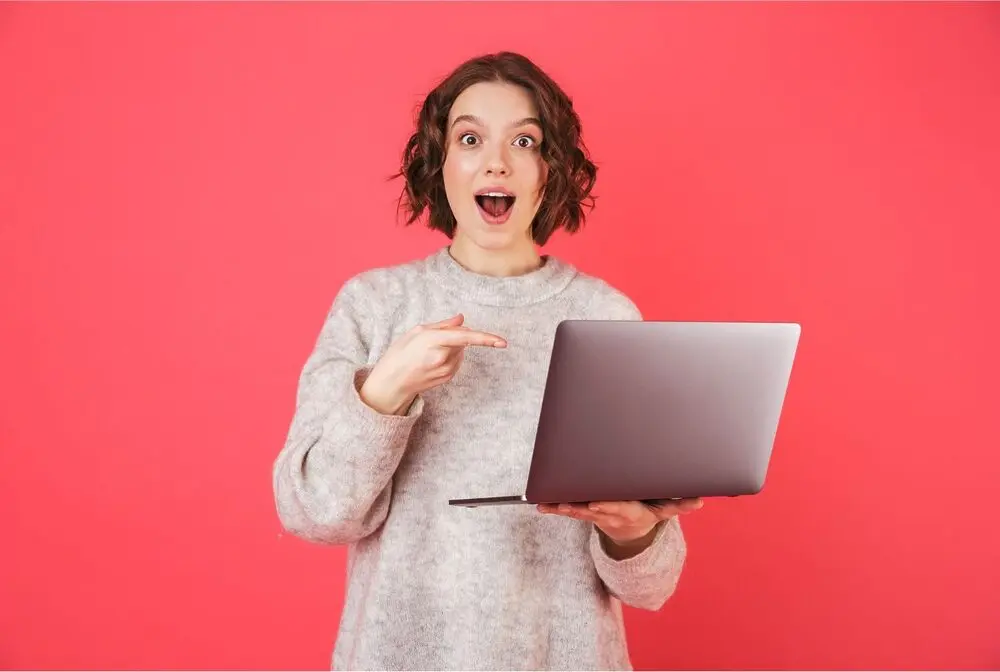 Short-haired woman smiling assertively while holding a laptop with one hand and pointing to it with the other one. She wears a grey sweater and has a blank reddish background behind her.
