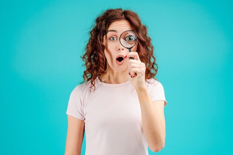 A woman with curly red hair holding a magnifying glass in front of a blue wall.
