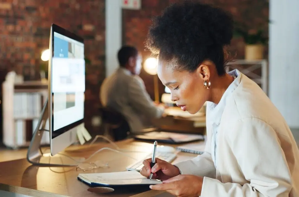 Elegant black woman at a coworker space taking handwritten notes on a notebook in front of a computer.