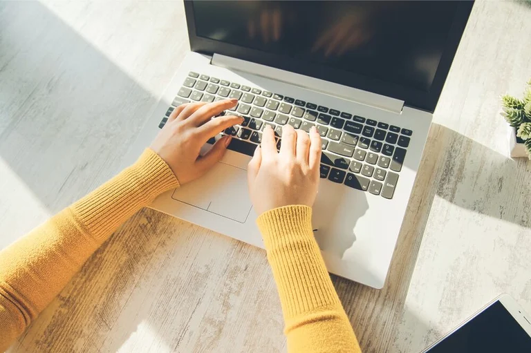 Hands typing on a laptop under morning sunlight.