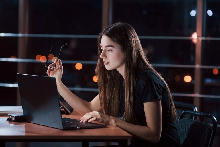 Long-haired woman wearing a black t-shirt and working with a black laptop on a modern rooftop at night.