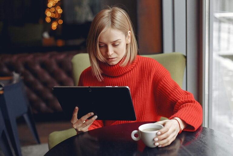 Short-haired blonde woman wearing a reddish sweater, holding a tablet with one hand and a cup of coffee with the other one.