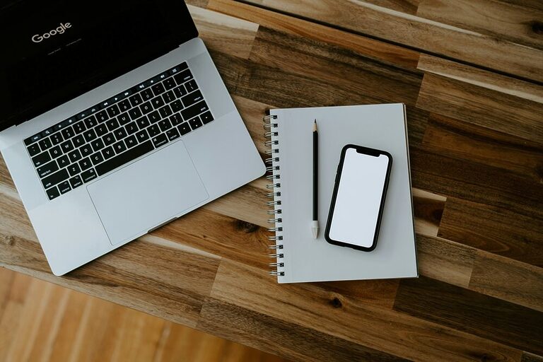 A spiral notebook, a smartphone, a pencil, and laptop on the table with Google on its screen.