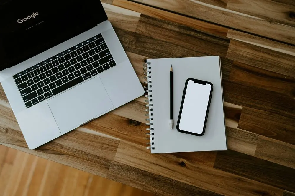A spiral notebook, a smartphone, a pencil, and laptop on the table with Google on its screen.