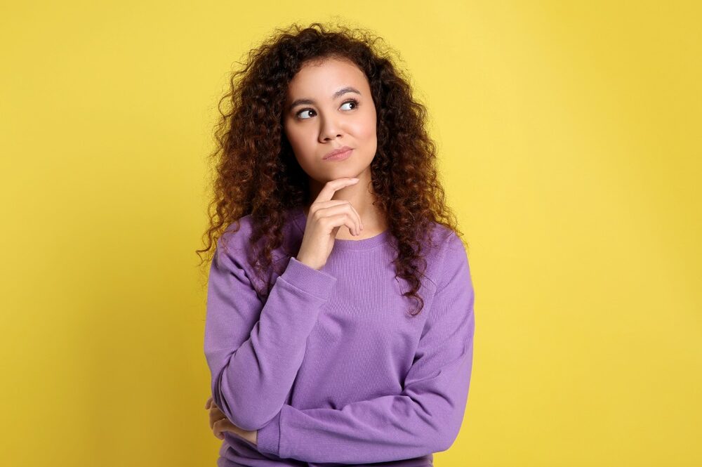 Black woman wearing a lilac long-sleeved t-shirt expressing doubt.