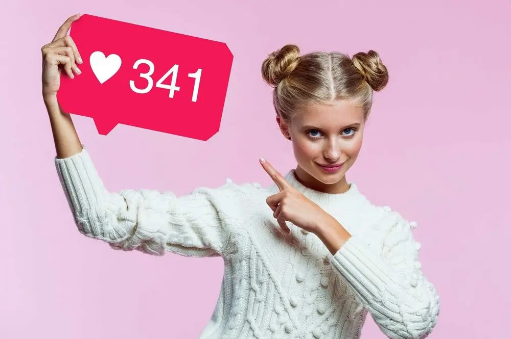 A blonde woman points with her left hand at a sign simulating the Instagram "like" button, which she holds in her right hand.