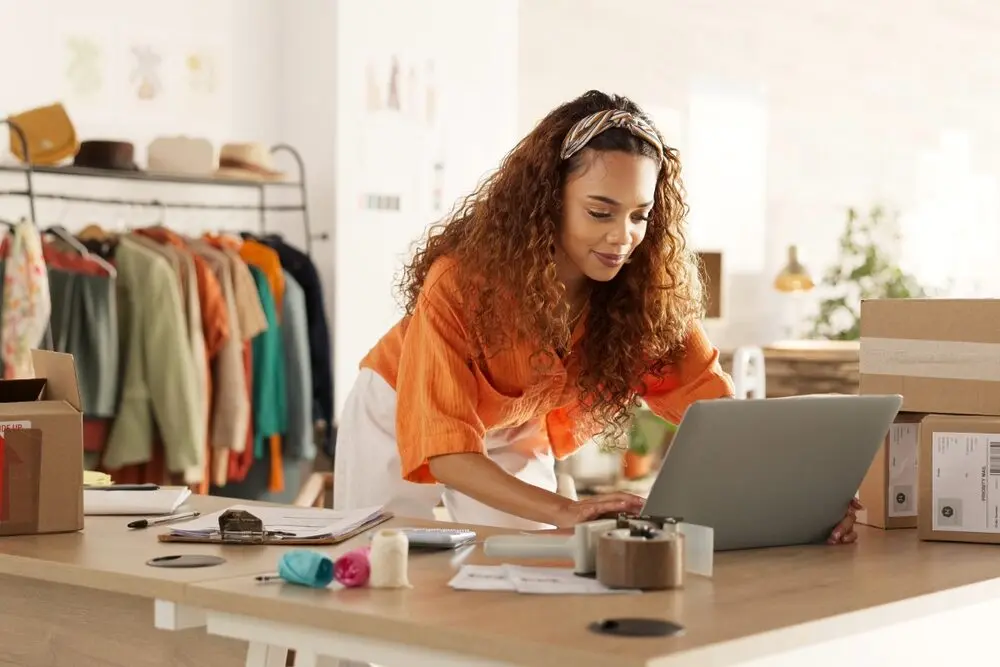 A woman leans over the counter to type something on the computer, with a clothing rack behind her.
