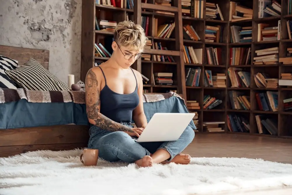 A woman with short hair is sitting on the floor of her room, leaning against the bed and working on a laptop.