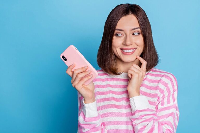 A woman with a bob haircut, in front of a light blue background, wearing a pink and white striped sweater and holding a cell phone in a pink case, smiling confidently, suggesting she has a good idea.