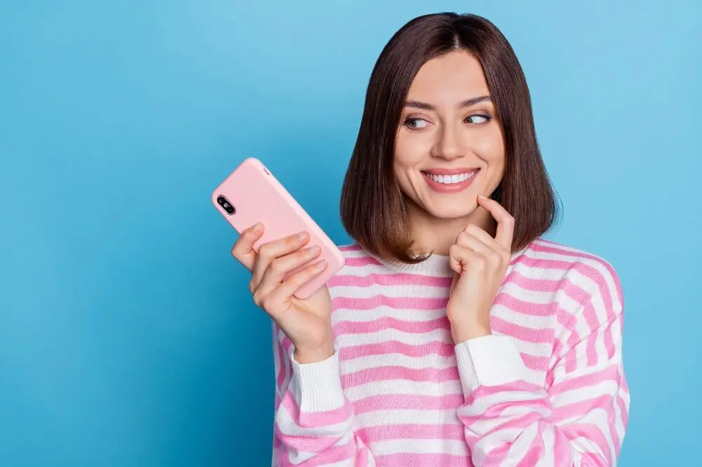 A woman with a bob haircut, in front of a light blue background, wearing a pink and white striped sweater and holding a cell phone in a pink case, smiling confidently, suggesting she has a good idea.