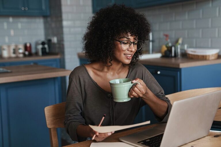 A black woman wearing glasses and holding a mug while reading something on her laptop. Behind her, there's a kitchen with blue furniture.