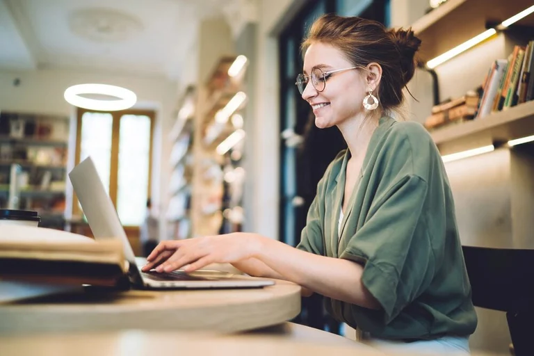 A woman wearing glasses and a green blouse smiles as she types on her laptop. She is in a coffee shop, sitting at a table in front of a bookcase.