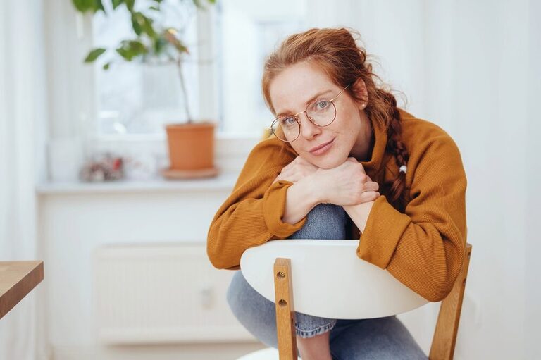 A red-haired woman sits facing the back of a chair, resting her hands and chin on her knee.