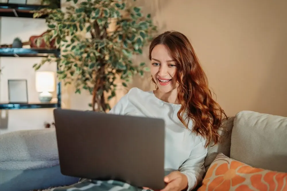 Red-haired woman comfortably sitting on a couch with a laptop above her legs.