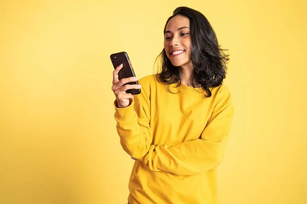 Dark-haired woman wearing a yellow hoodie and smiling to a smartphone screen that she's holding with her right hand in front of a yellow background.