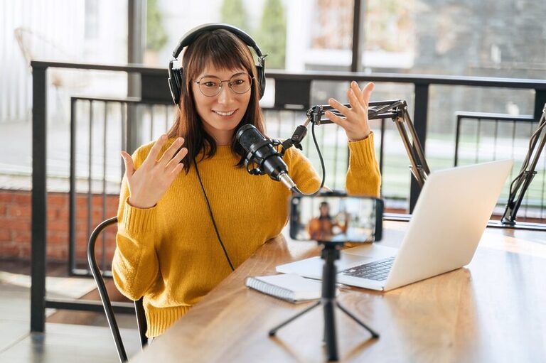 A content creator is recording a video with a smartphone propped up on a table. She is wearing a yellow sweater and headphones.