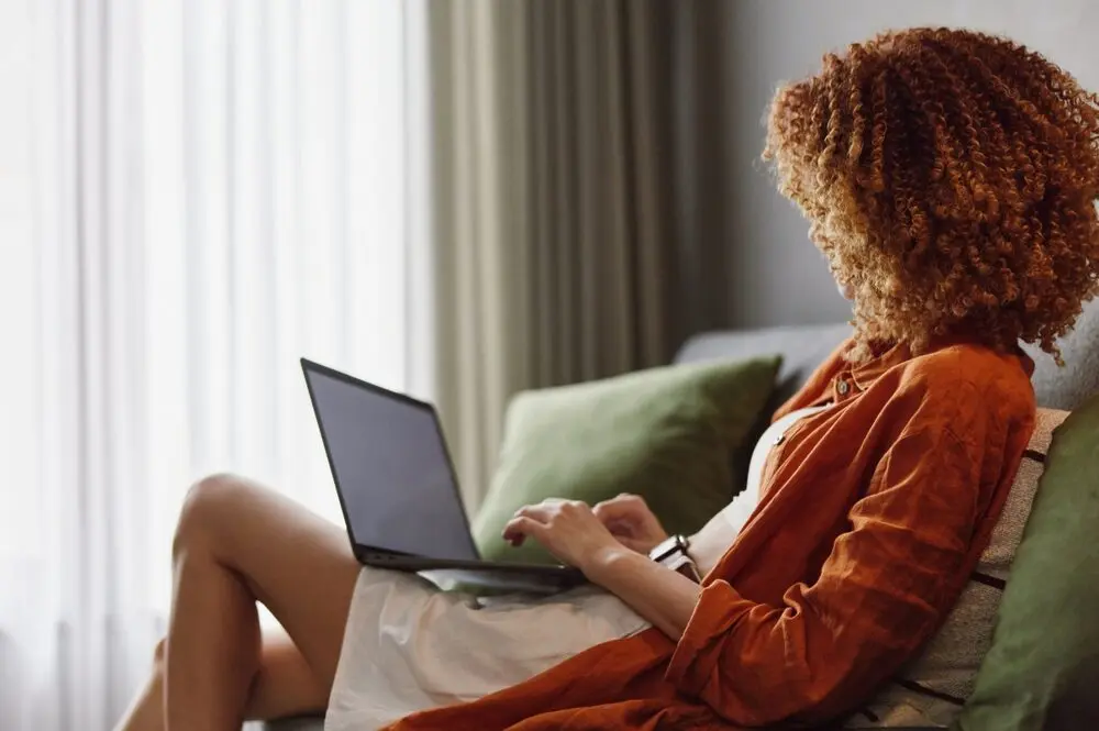 Black woman wearing an orange carding and sitting on a couch while a laptop rests on her tights.