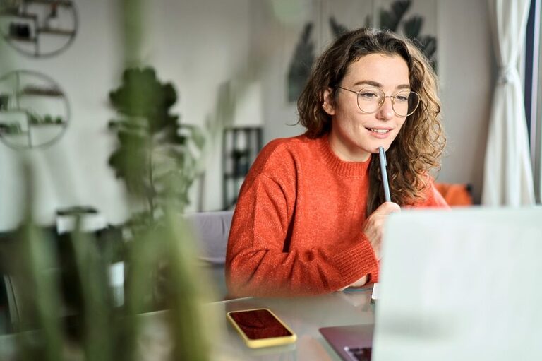 A woman with brown hair, wearing glasses and an orange sweater, presses a pen against the tip of her lower lip, and smiles calmly but excitedly while looking at a laptop screen.